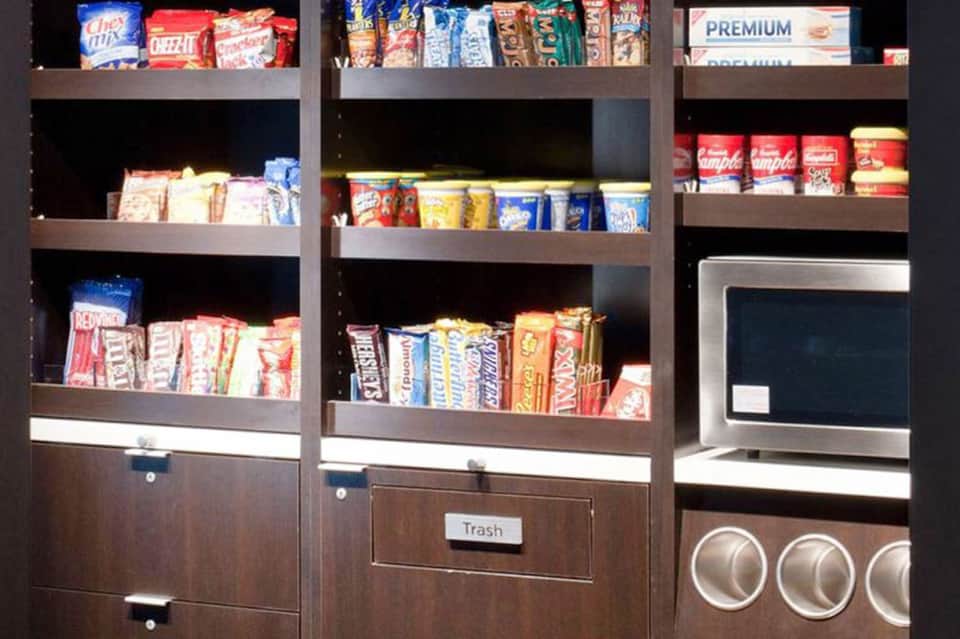 Vending machine shelves stocked with snacks, beverages, and packaged goods with trash bin below