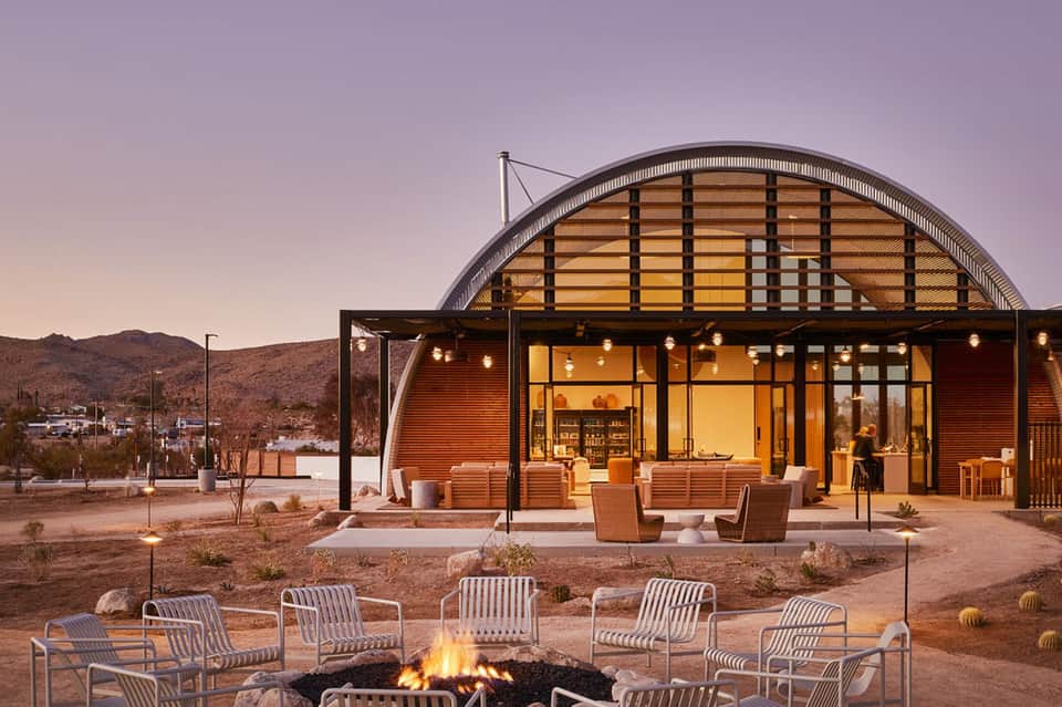 Desert resort pavilion at dusk with curved architectural design, firepit seating area, warm lighting, and desert mountains