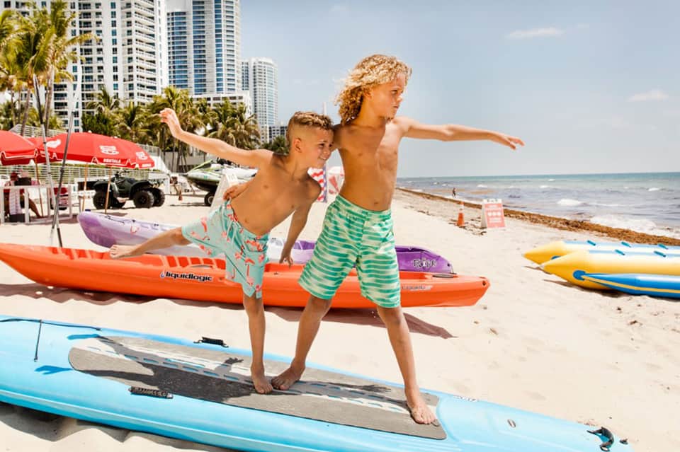 Two children in swim trunks balancing on surfboards on beach with high-rise hotels
