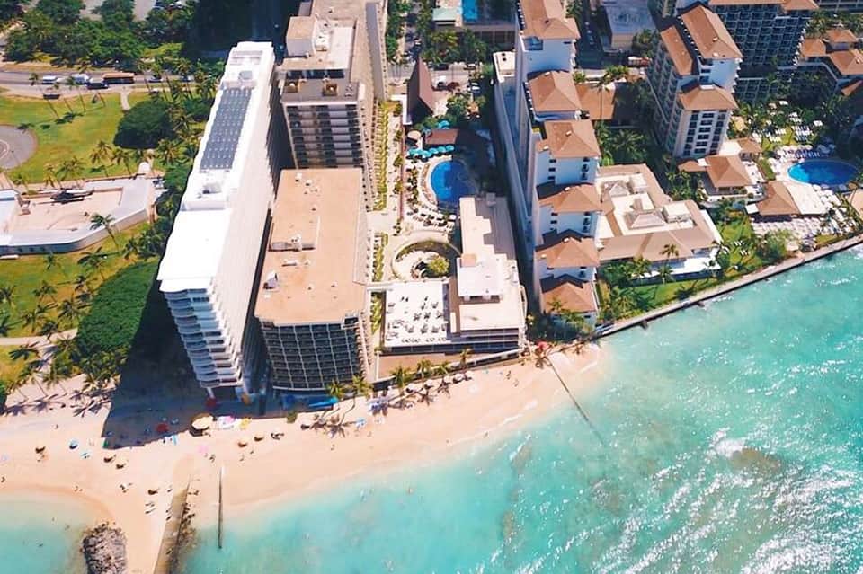 Aerial view of beachfront resort with modern buildings, turquoise ocean water, and sandy beach with loungers