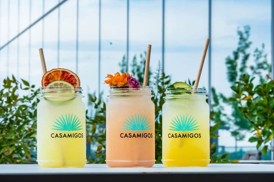 Three colorful Casamigos cocktails with citrus garnishes and flowers against ocean view