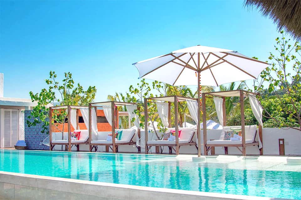 Resort poolside with turquoise water, white umbrellas, cabanas with flowing curtains, and tropical vegetation