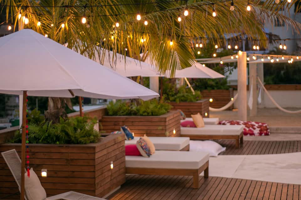 Rooftop terrace at dusk with loungers, white umbrellas, string lights, and palm fronds overhead
