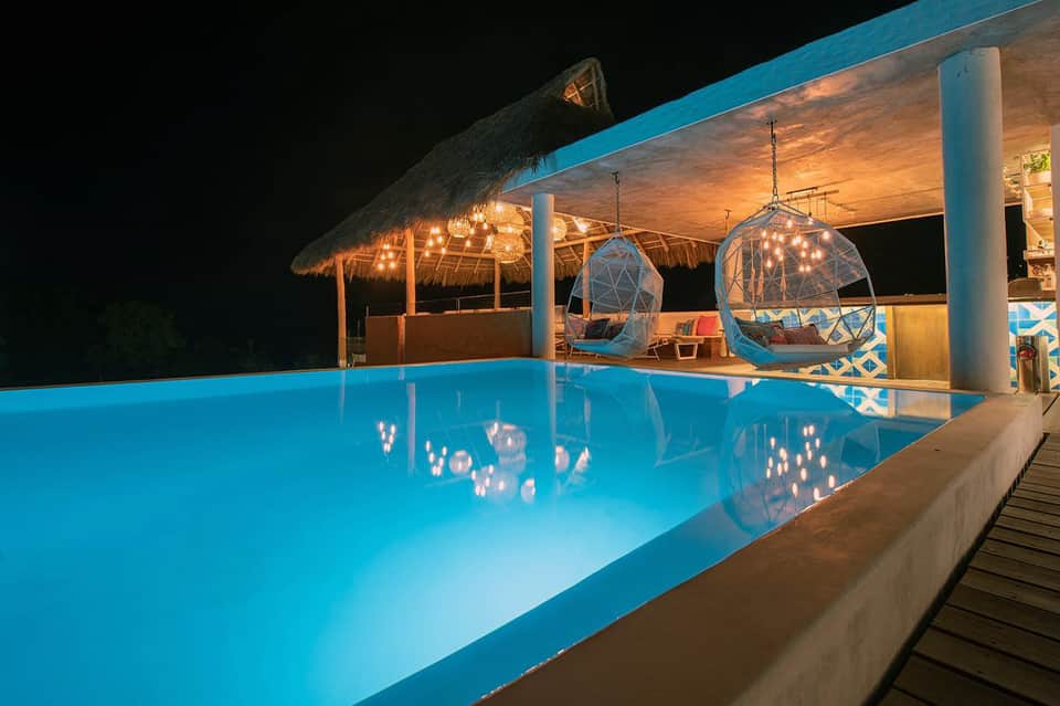 Illuminated outdoor pool at night with thatched-roof structure and hanging bubble chairs