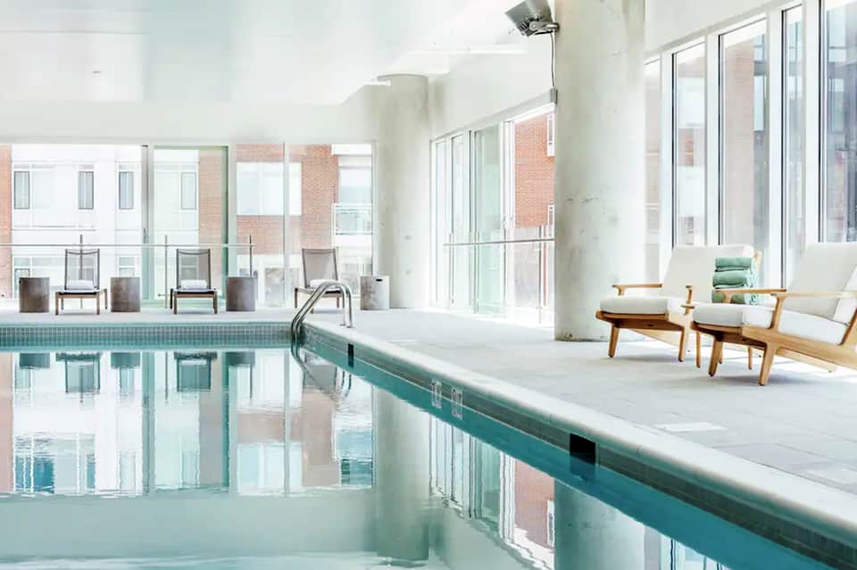 Indoor swimming pool with teal edge, wooden lounge chairs, and floor-to-ceiling windows
