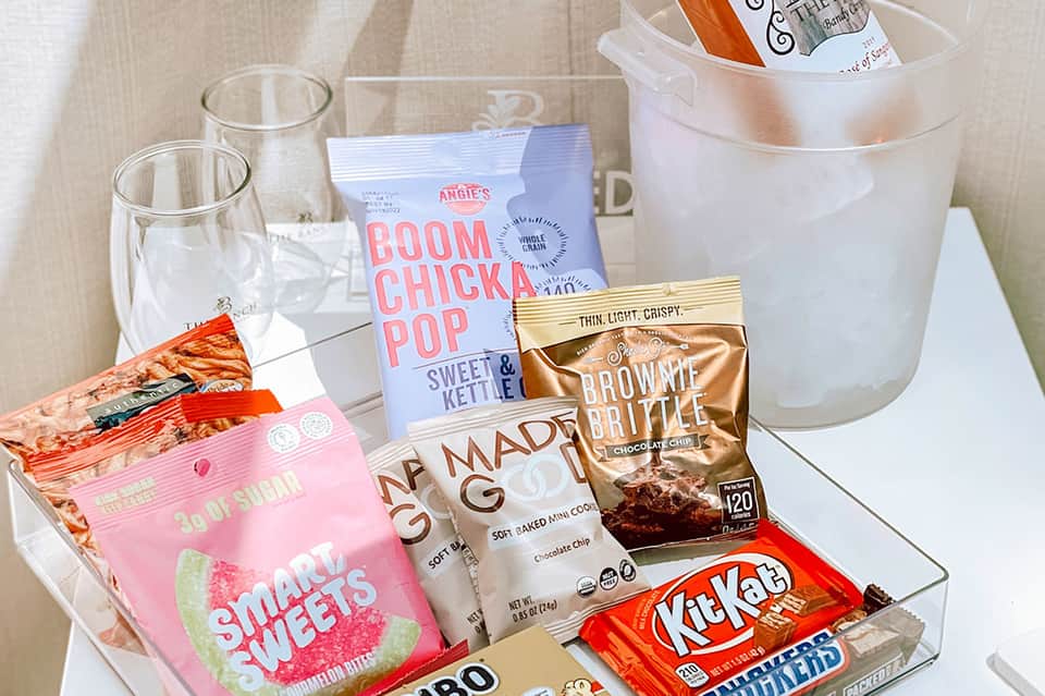 In-room mini bar with assorted snacks, candy, drinks, and welcome amenities on white tray
