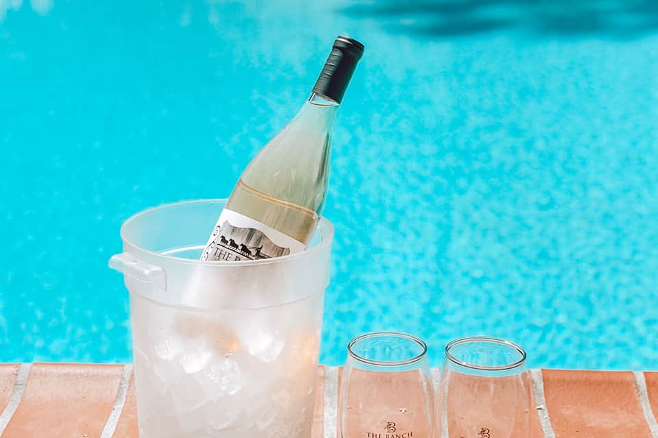 Wine bottle chilling in ice bucket by turquoise pool water with two glasses
