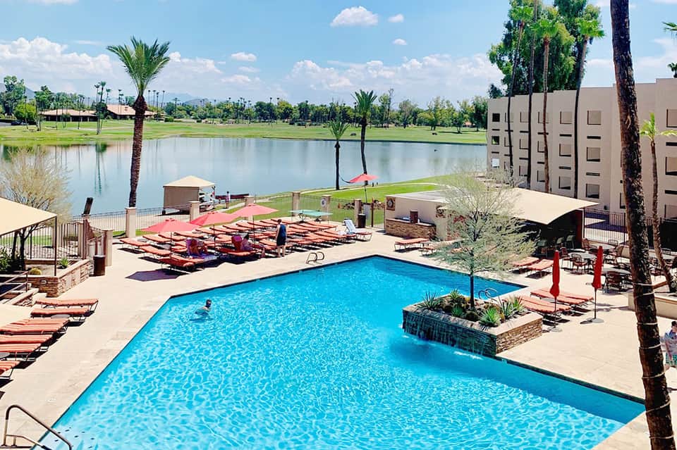 Resort pool area with lounge chairs overlooking lake and palm trees under blue sky