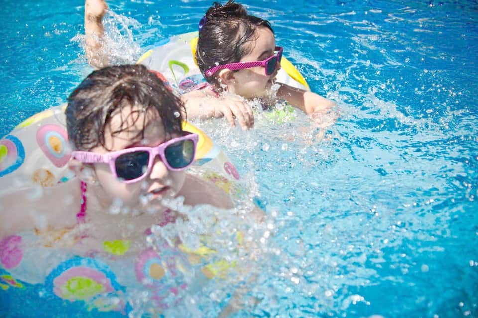 Children swimming in bright blue pool wearing colorful floaties and sunglasses
