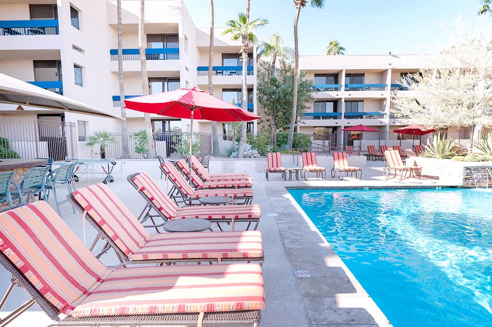 Resort pool area with pink and white striped loungers, red umbrellas, and multi-story white buildings with blue railings