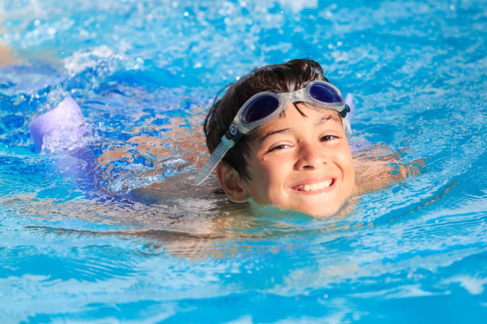 Young boy swimming in clear blue pool wearing goggles and smiling