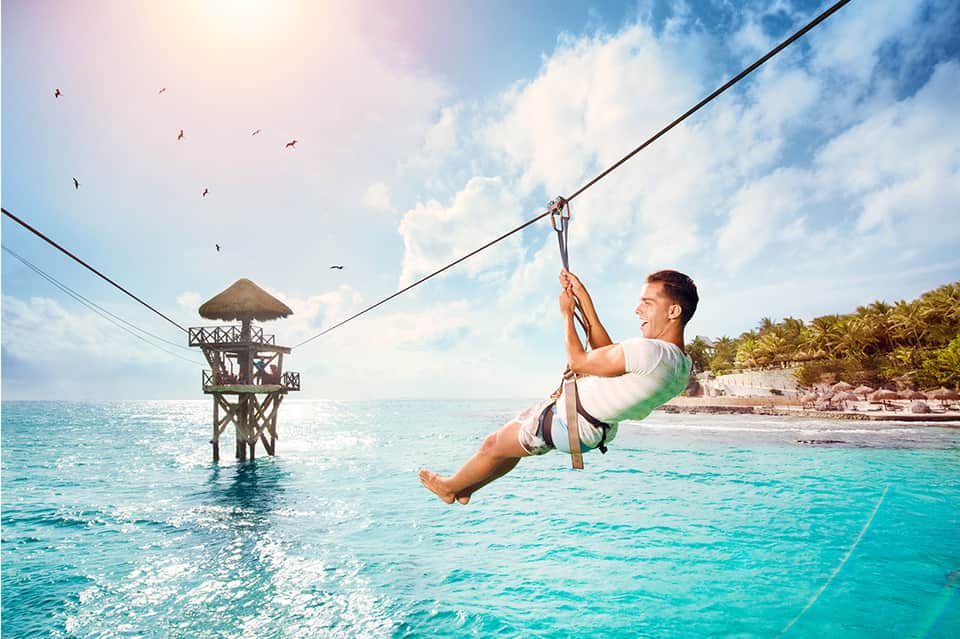 Man zip-lining over turquoise ocean water toward a beach platform with palm trees
