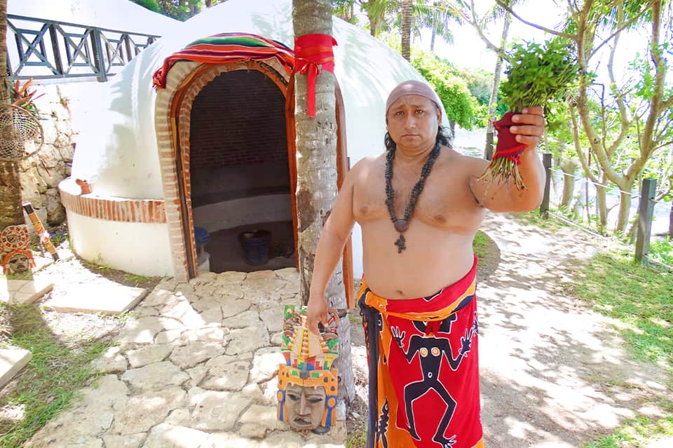 Indigenous man in traditional red ceremonial dress holding botanical materials outside tiki hut