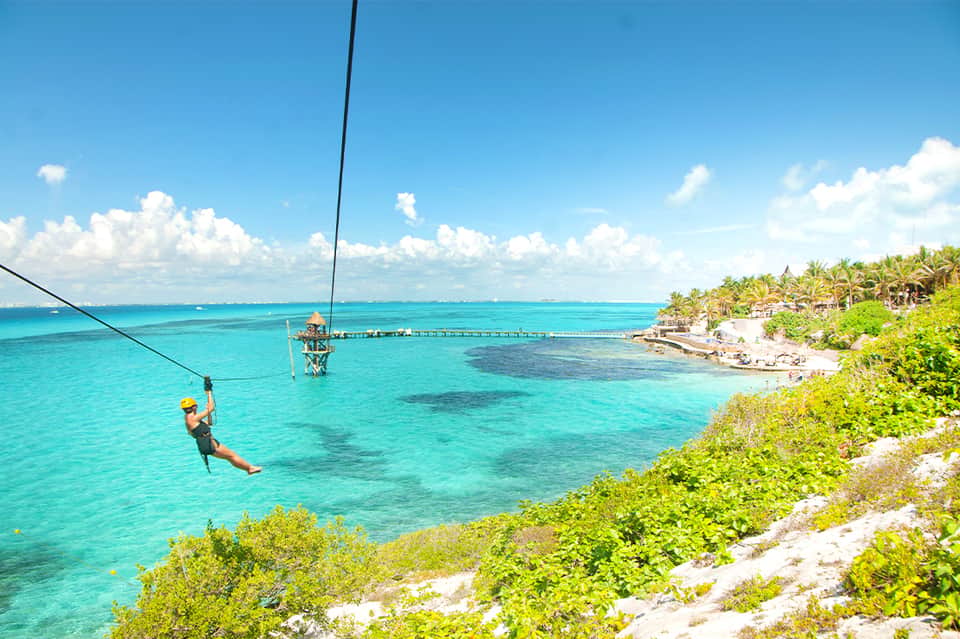 Person zip-lining over turquoise ocean water with palm trees and beach resort visible below