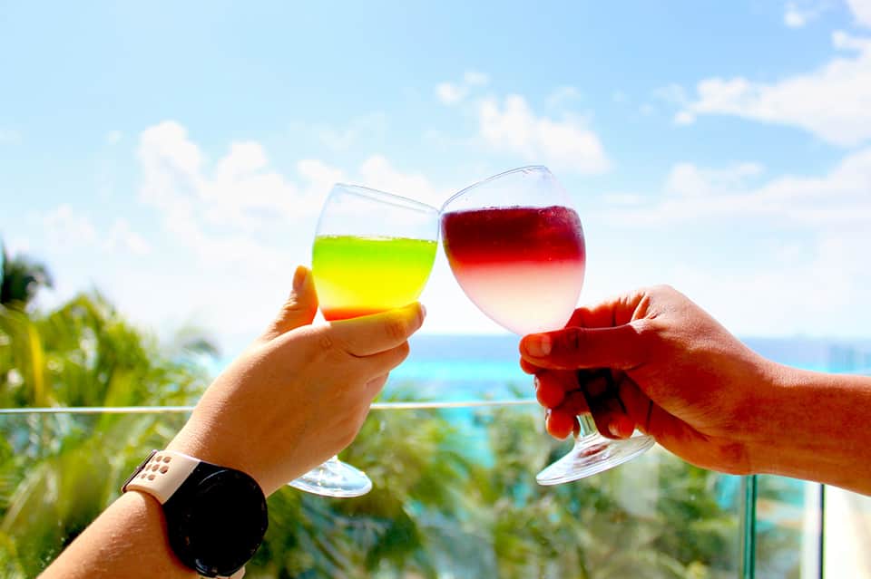 Two hands toasting with colorful cocktails overlooking tropical ocean view