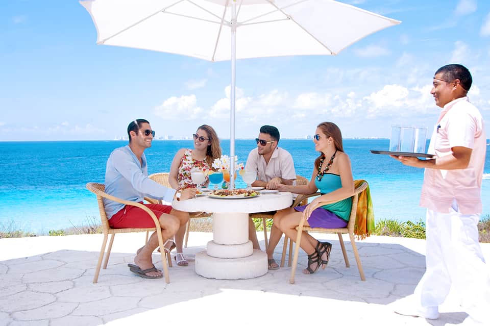 Group dining at beachfront table with umbrella, server in pink uniform, turquoise ocean and sky backdrop