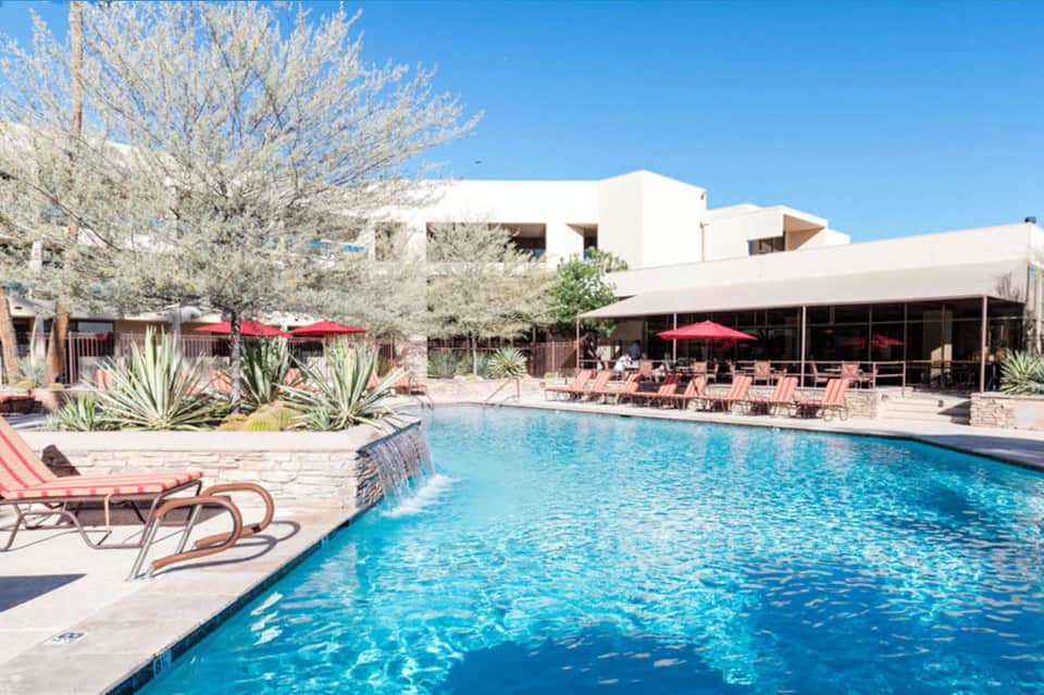Resort pool with crystal clear turquoise water, red umbrellas, lounge chairs, and modern white architecture.
