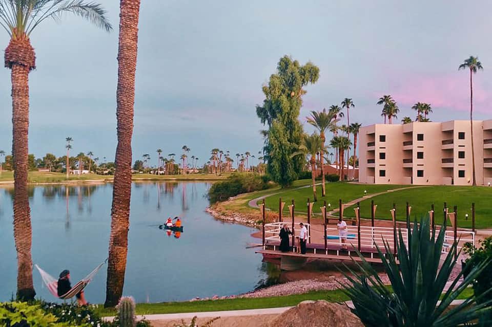 Resort grounds at dusk with pink building, palm trees, lagoon, dock, and person in hammock