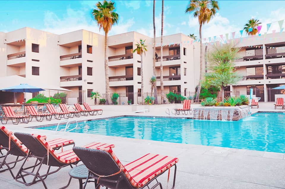 Resort pool area with colorful striped lounge chairs, palm trees, and white multi-story hotel building backdrop