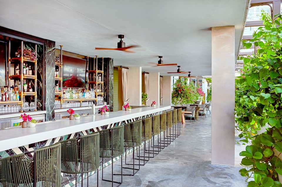 Modern bar with decorative wall backdrop, bar seating, ceiling fans, and tropical plants