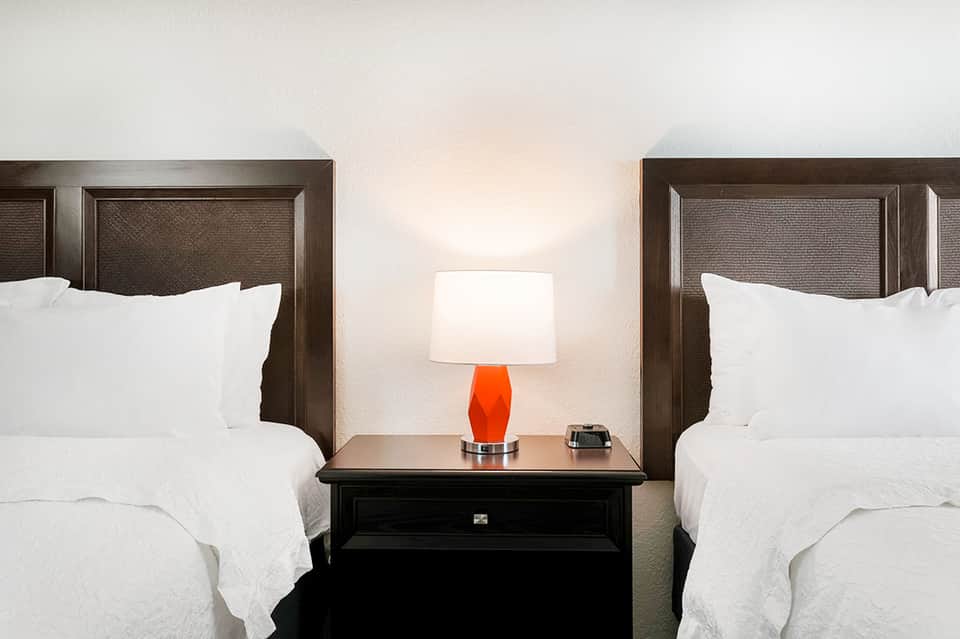 Modern hotel bedroom with two queen beds, dark wooden headboards, white bedding, and orange table lamp