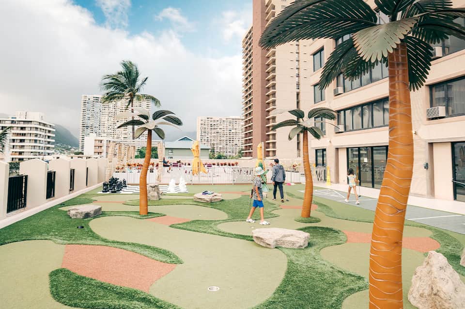 Outdoor mini golf course with synthetic turf, palm trees, and high-rise buildings in background