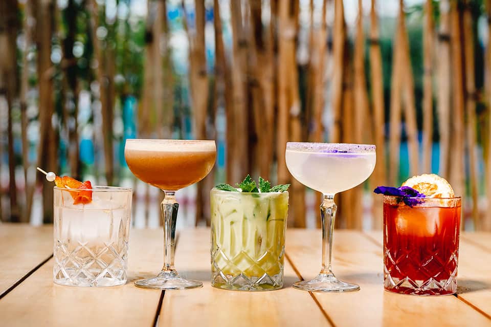 Five colorful cocktails on wooden deck with beach fence background and tropical garnishes