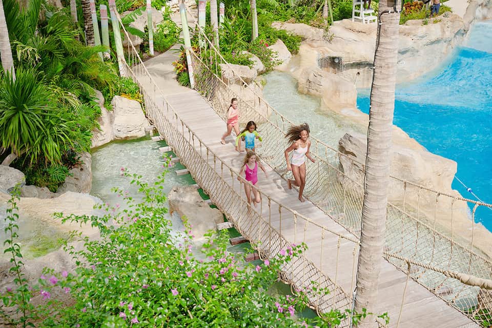 Children walking across rope bridge over turquoise water surrounded by tropical vegetation