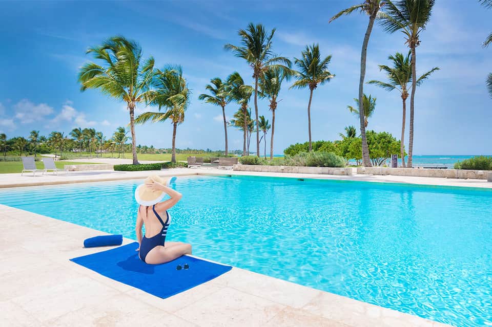 Woman in hat sitting poolside with palm trees and ocean beyond