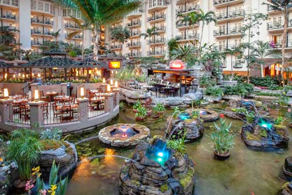 Indoor atrium with dining areas, koi pond with lit rocks, tropical plants, and multi-story hotel buildings surrounding the space