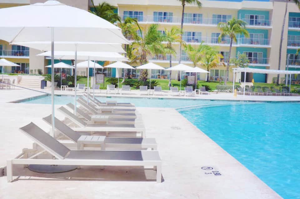 Resort pool with white lounge chairs and umbrellas, flanked by palm trees and colorful building