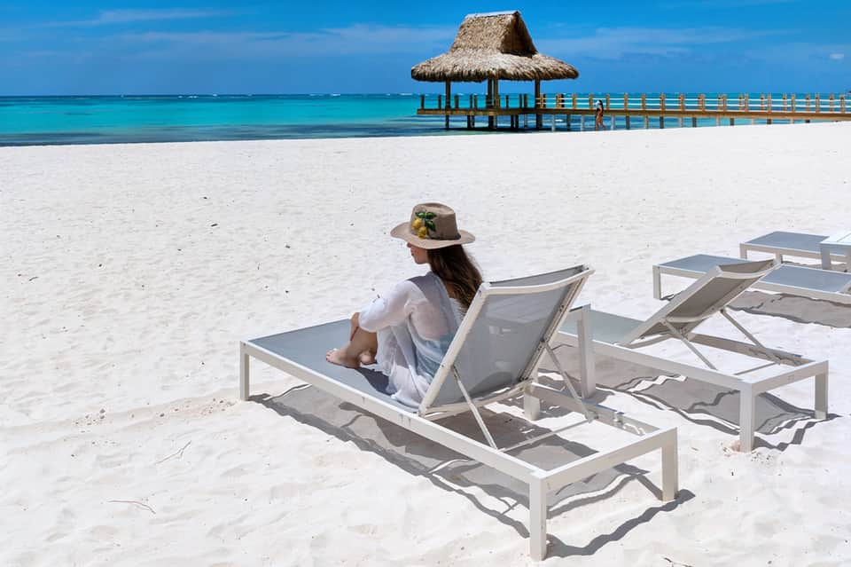 Woman relaxing on beach lounge chair facing turquoise ocean with thatched pier structure