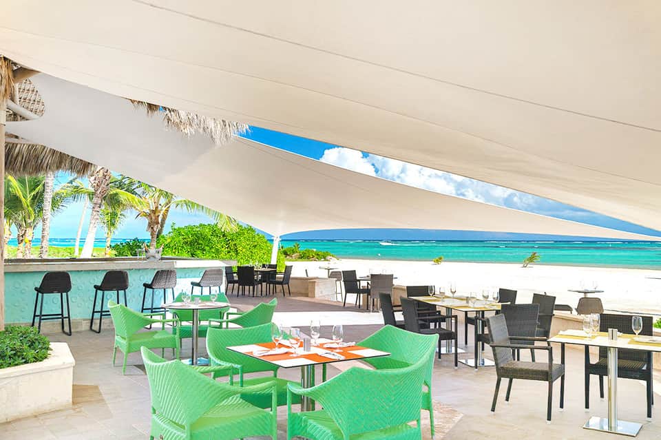 Beachfront restaurant terrace with green chairs, white shade sail, turquoise ocean view, and tropical dining ambiance