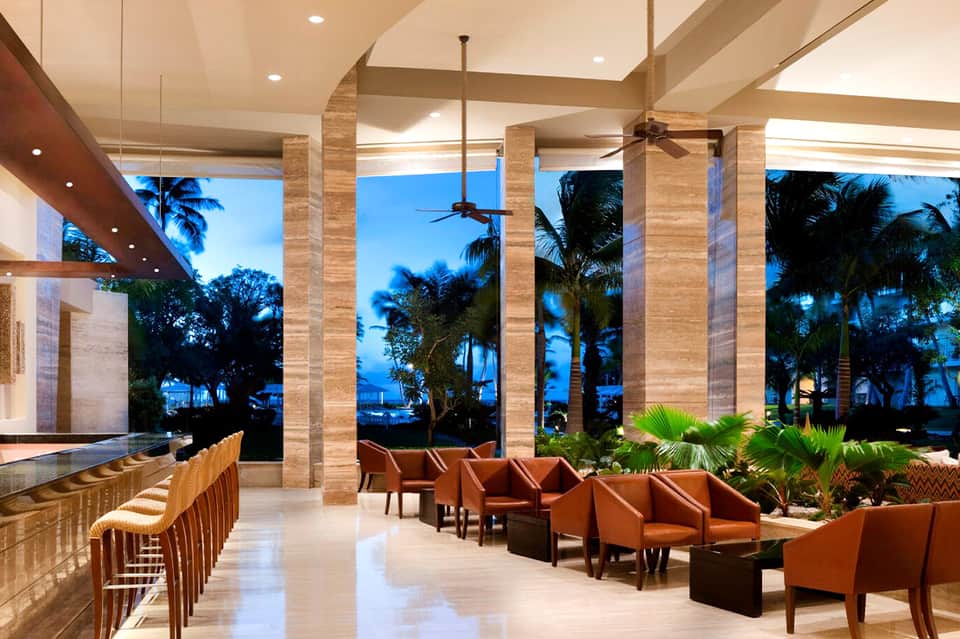 Luxury resort lounge at dusk with travertine columns, ocean views, palm trees, and burnt orange seating