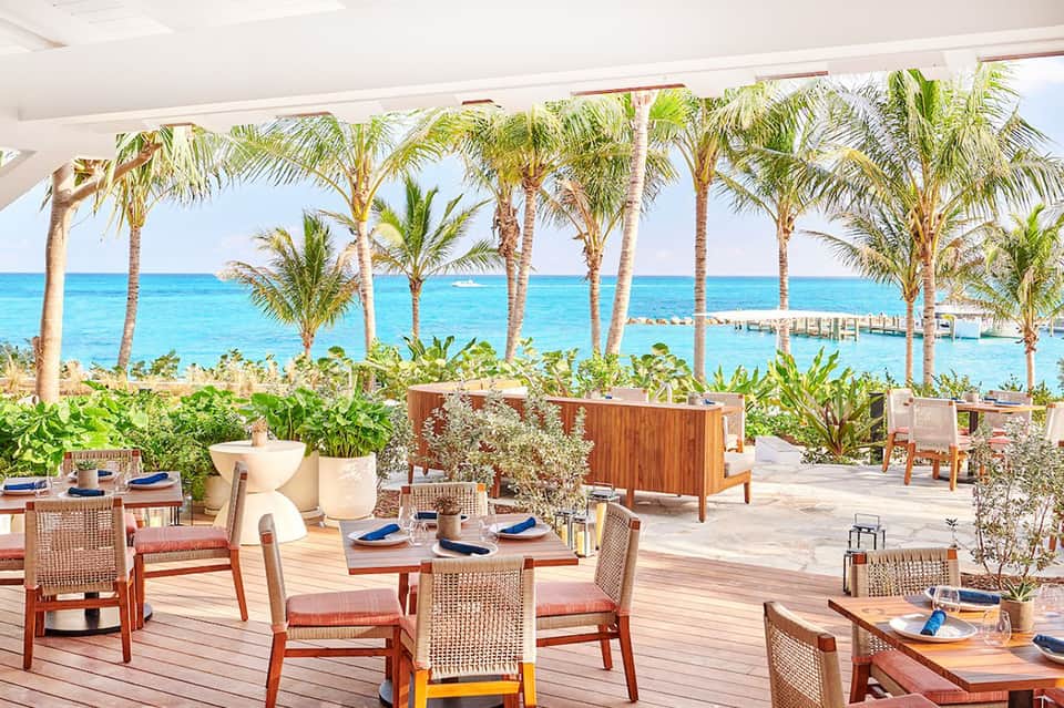 Beachfront restaurant patio with wooden dining tables, rattan chairs, palm trees, and ocean view at horizon