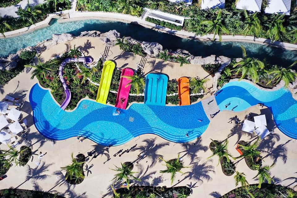 Aerial view of wave-shaped swimming pool with colorful water slides surrounded by palm trees and sand