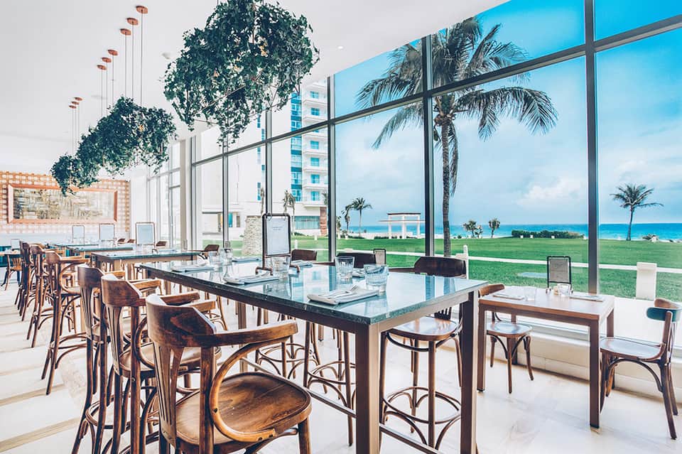 Modern restaurant interior with floor-to-ceiling windows overlooking tropical beach and palm trees