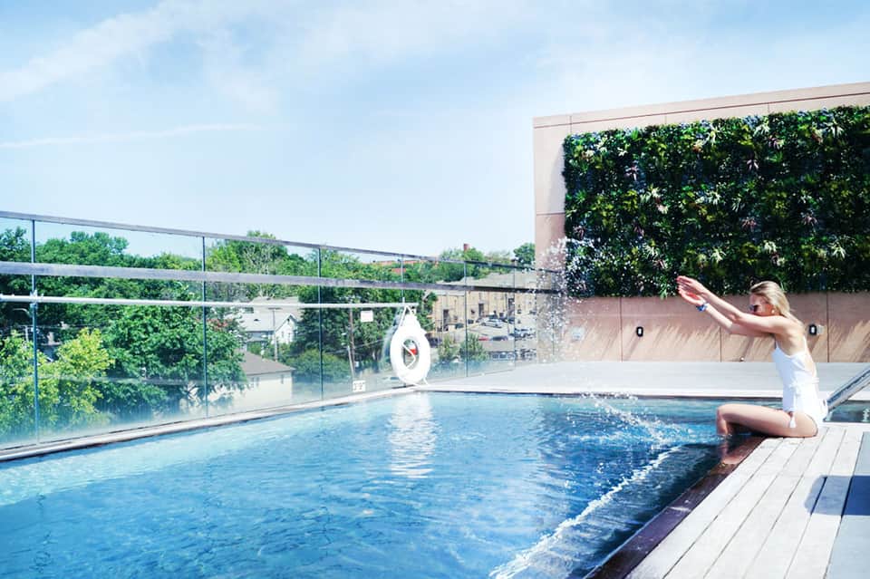 Woman sitting poolside in white outfit at rooftop pool with glass railings and green plant wall