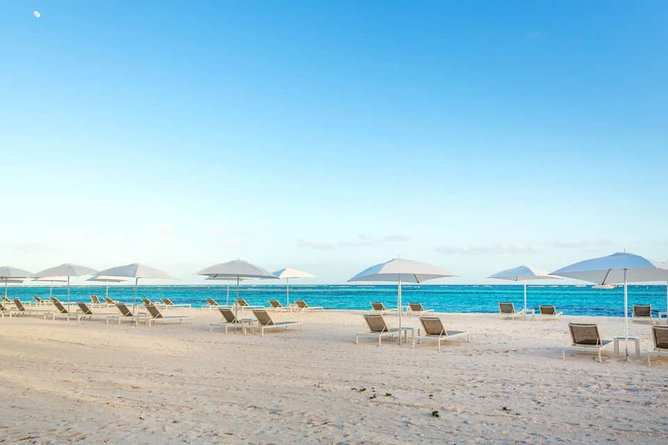 Wide sandy beach with lounge chairs, white umbrellas, and turquoise ocean under blue sky