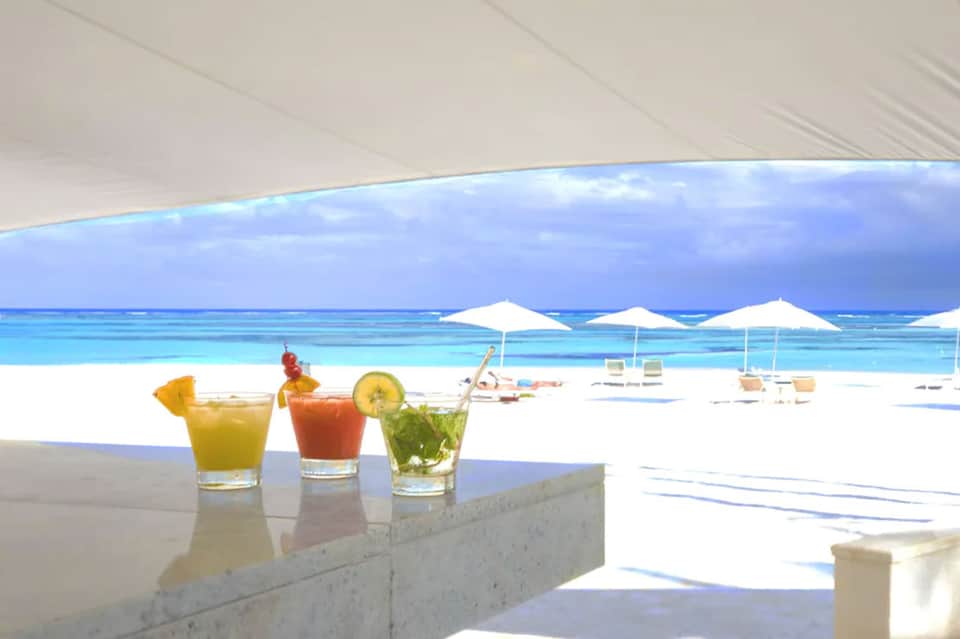 Three tropical cocktails on marble counter with beach, umbrellas, and turquoise water in background
