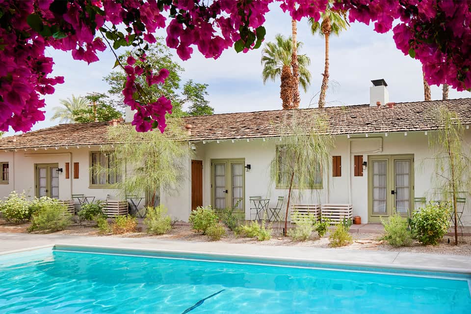 White mid-century resort building with magenta bougainvillea overhead, turquoise pool in foreground, palm trees
