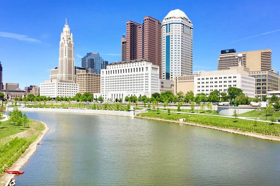 Urban skyline featuring historic tower and modern buildings reflected in river with green park