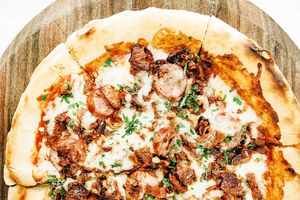 Wood-fired pizza topped with meat, cheese, and fresh herbs on wooden serving board