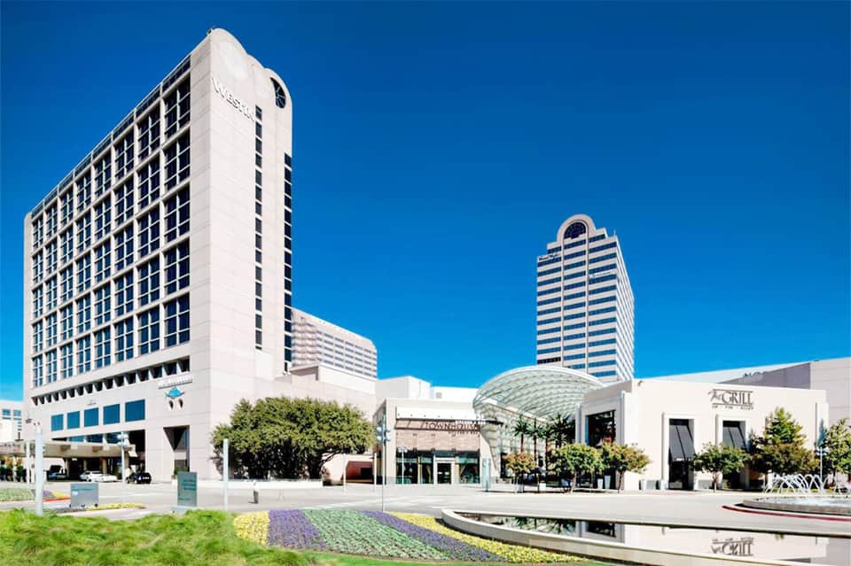 Modern hotel exterior with white high-rise towers and contemporary plaza featuring landscaped gardens and reflecting pool