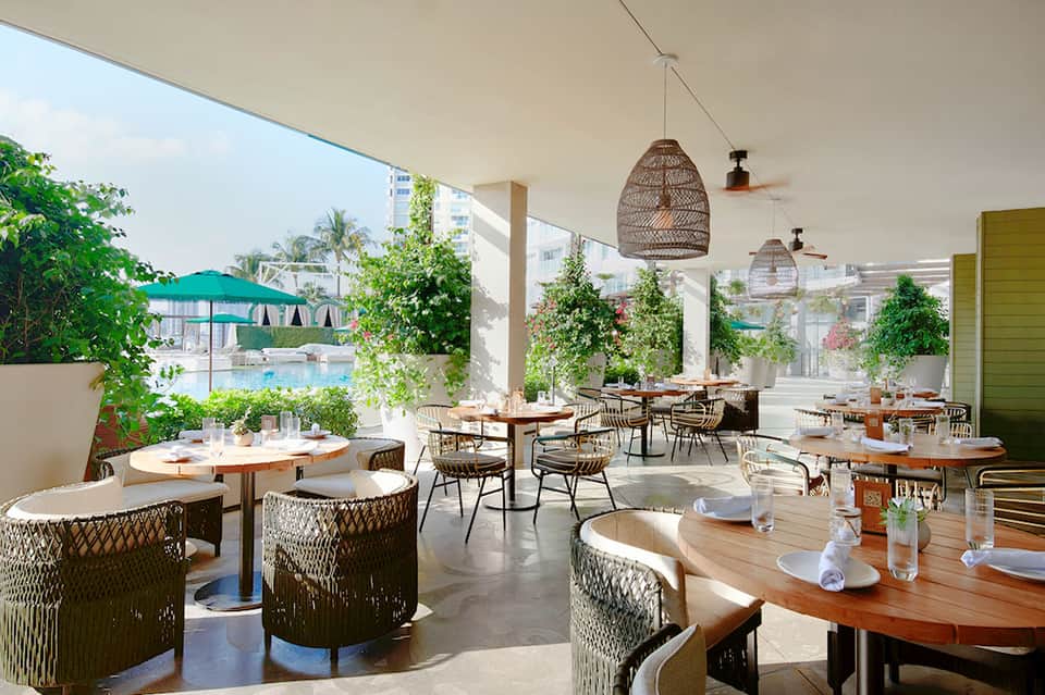 Modern outdoor restaurant terrace with woven pendant lights, wooden tables, and pool view