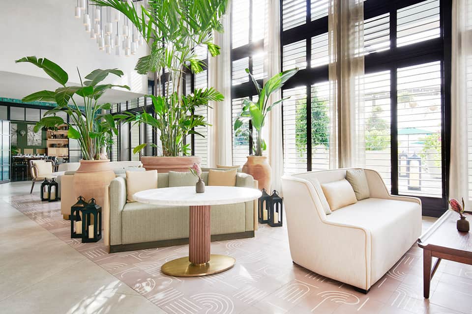 Bright modern lobby lounge with cream seating, tall potted plants, natural light from black-framed windows