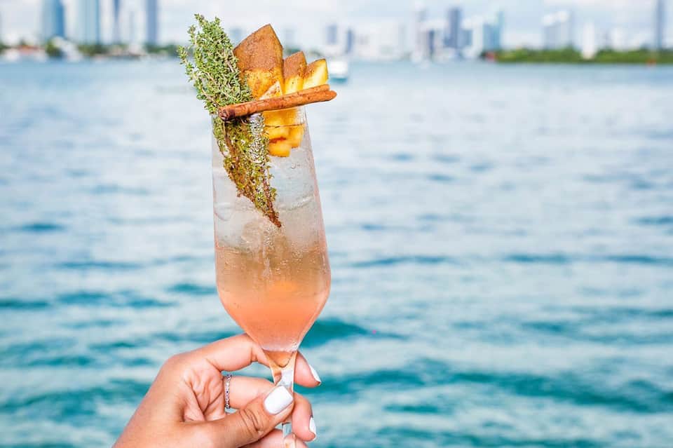 Tropical cocktail with fresh herbs and gold leaf garnish held over blue ocean water with city skyline backdrop