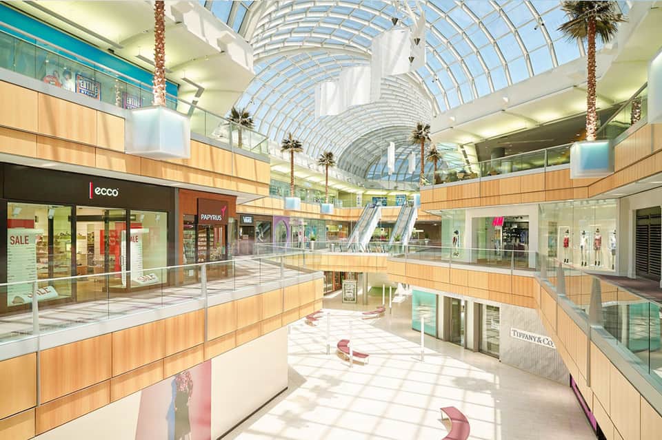 Modern mall interior with glass arched ceiling, palm trees, and retail storefronts