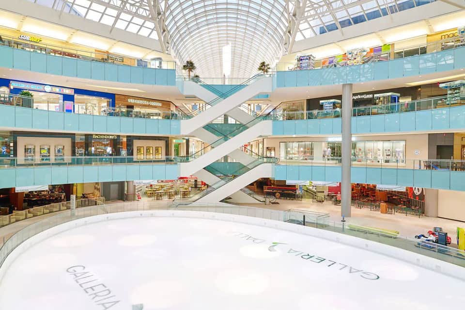 Multi-level shopping mall interior with escalators, glass railings, and modern architectural design