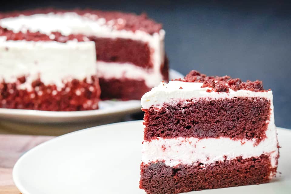 Slice of red velvet cake with white frosting on white plate with whole cake in background
